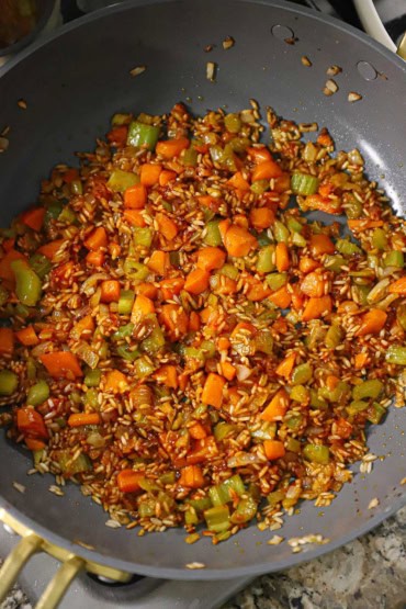 An overhead view of a large deep skillet filled with sautéed pieces of carrot, celery, and onion mixed with long-grain white rice and tomato paste.