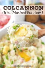 A close-up view of a wooden spoon being used to hold up a large serving of colcannon (Irish mashed potatoes) with a pad of melting butter on top.