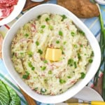 An overhead view of a large bowl filled colcannon (Irish mashed potatoes) with a couple tabs of butter in the center and whole russet potatoes resting nearby.