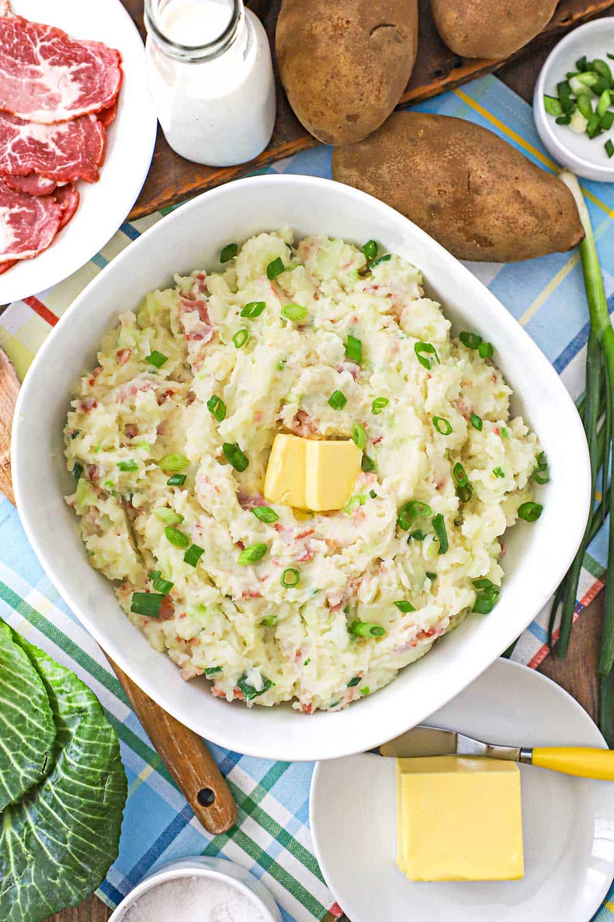 An overhead view of a large bowl filled colcannon (Irish mashed potatoes) with a couple tabs of butter in the center and whole russet potatoes resting nearby.
