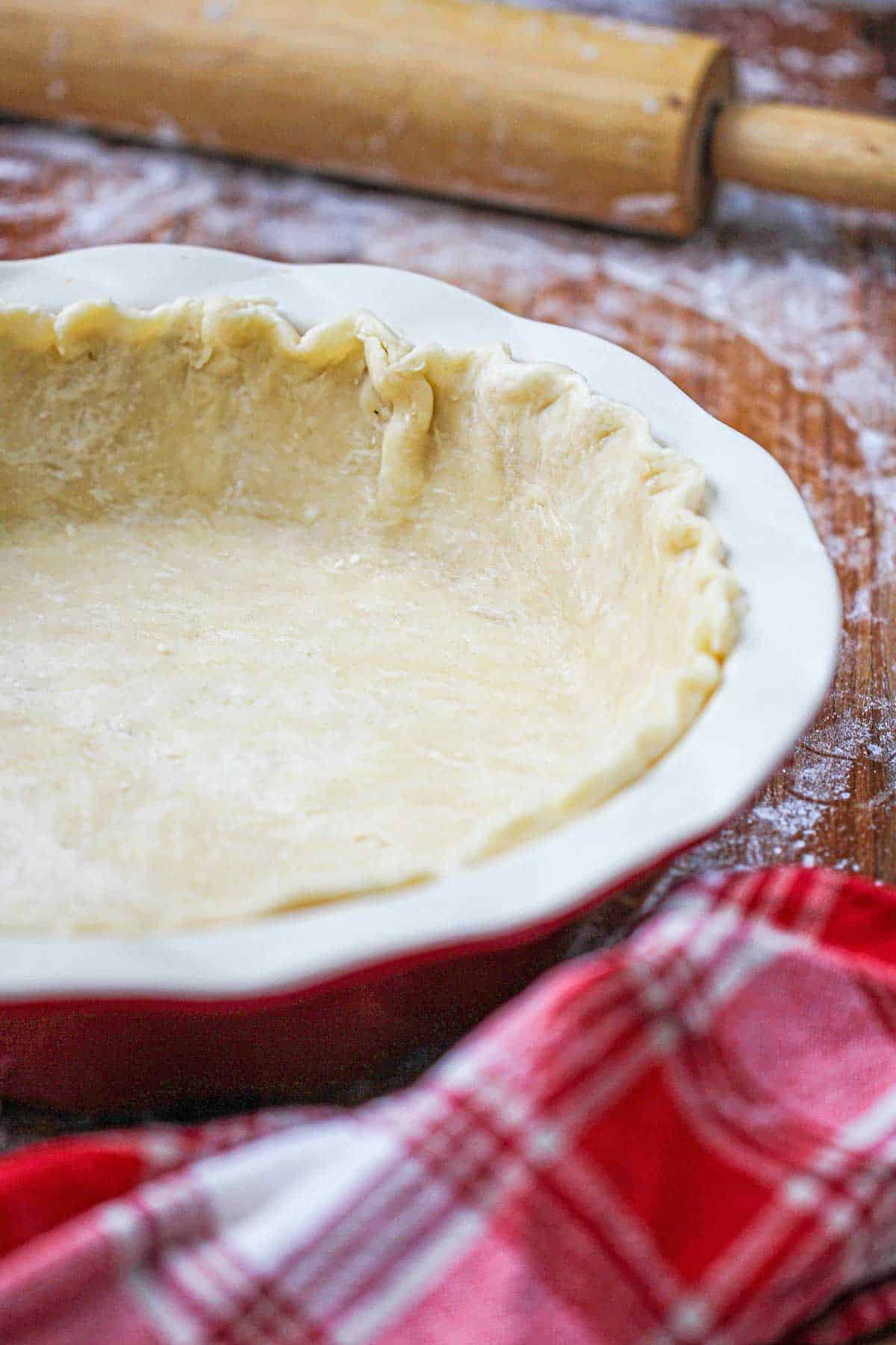 Easy pie crust dough that has been fitted into a ceramic pie dish with a floured rolling pin nearby. 