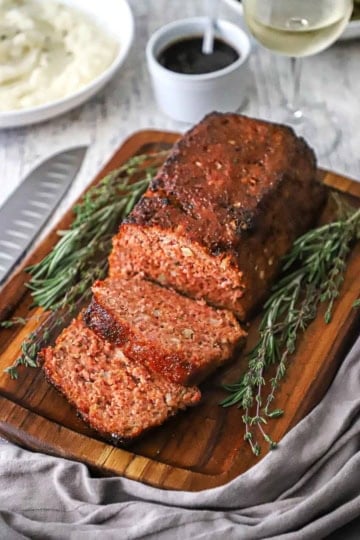 A glazed ham loaf sitting on a small cutting board and has a few slices cut sitting next to sprigs of thyme and rosemary.