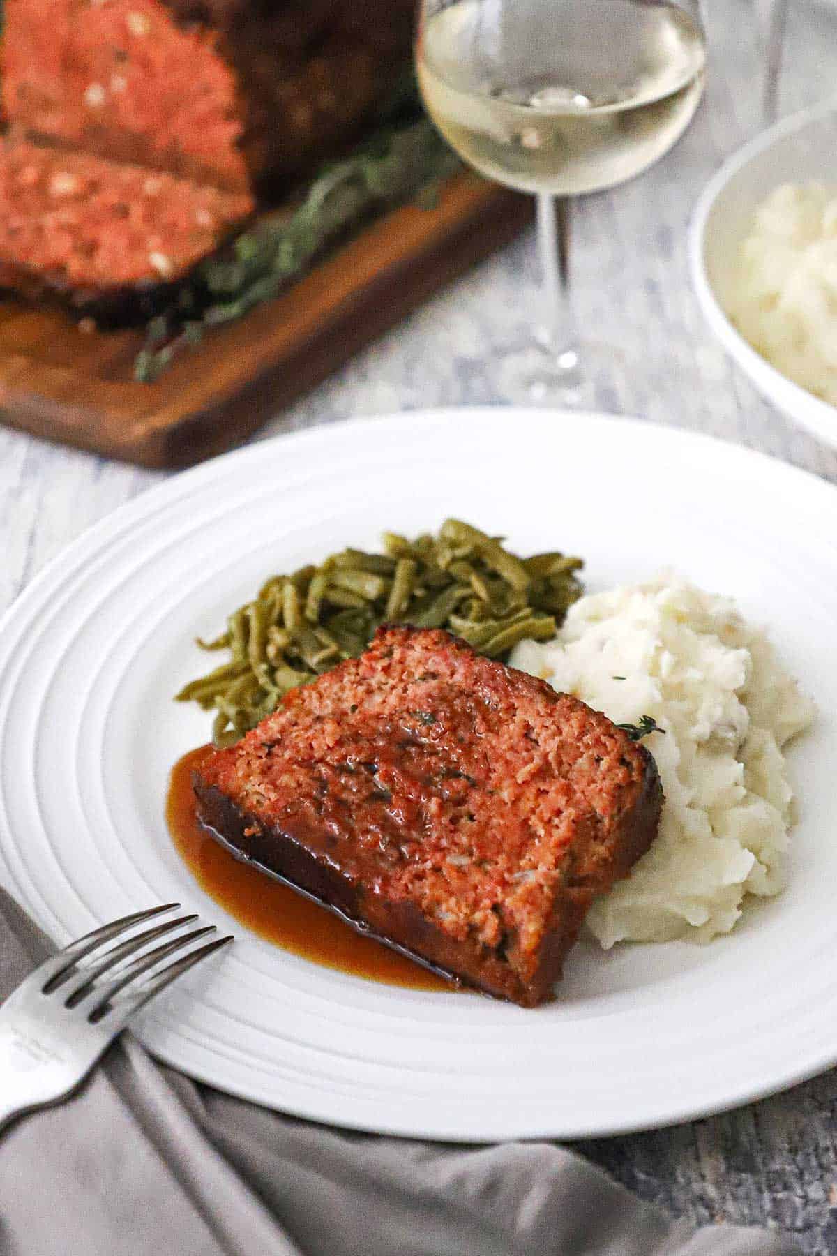 A white dinner plate filled with a serving of  slized glazed ham loaf on a pile of mashed potatoes next to Southern-style green beans. 