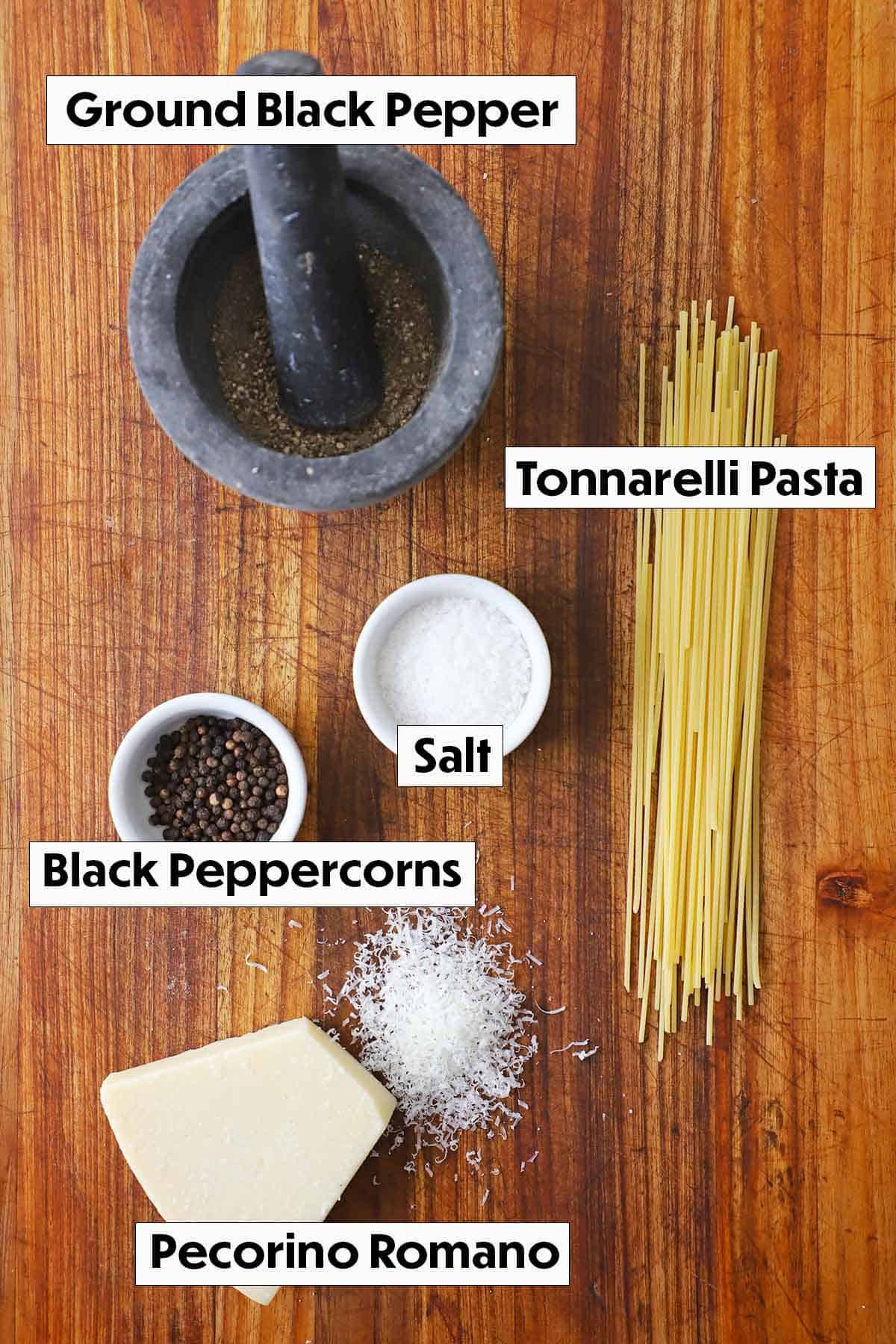 An arrangement of ingredients for cacio e pepe including a mortar and pestle filled with ground black pepper, dried pasta, salt, and a block of pecorino romano all on a wooden cutting board  