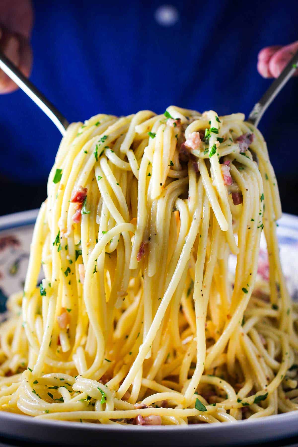 A person using a two serving utensils to lift up a large amount of pasta carbonara from a large shallow pasta bowl. 