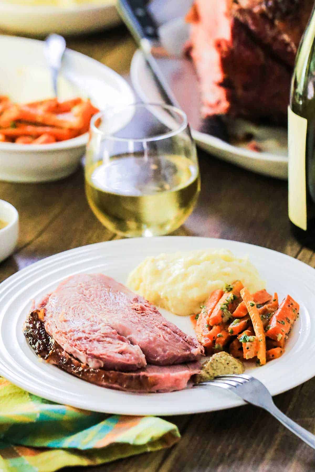 A white dinner plate filled with several slices of honey bourbon glaze ham next to braised carrots, cooked potatoes and a glass of white wine nearby. 