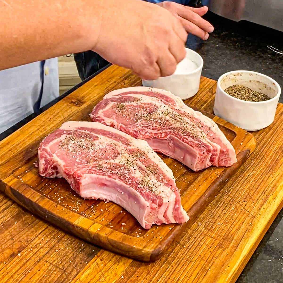 A person sprinkling salt and pepper over two uncooked pork chops on a cutting board. 