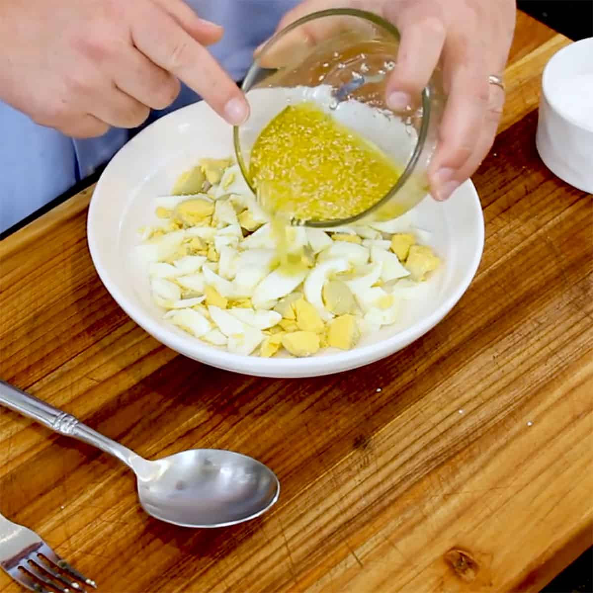 A person pouring a lemon sesame vinaigrette over chopped hard boiled eggs in a shallow white bowl. 