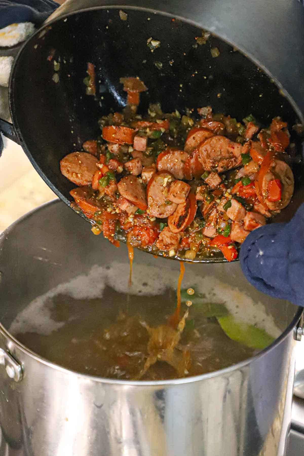 A person dumping a black pot filled with sautéed sausage slices and cooked vegetables and seasonings into a large stock pot filled with simmering broth.