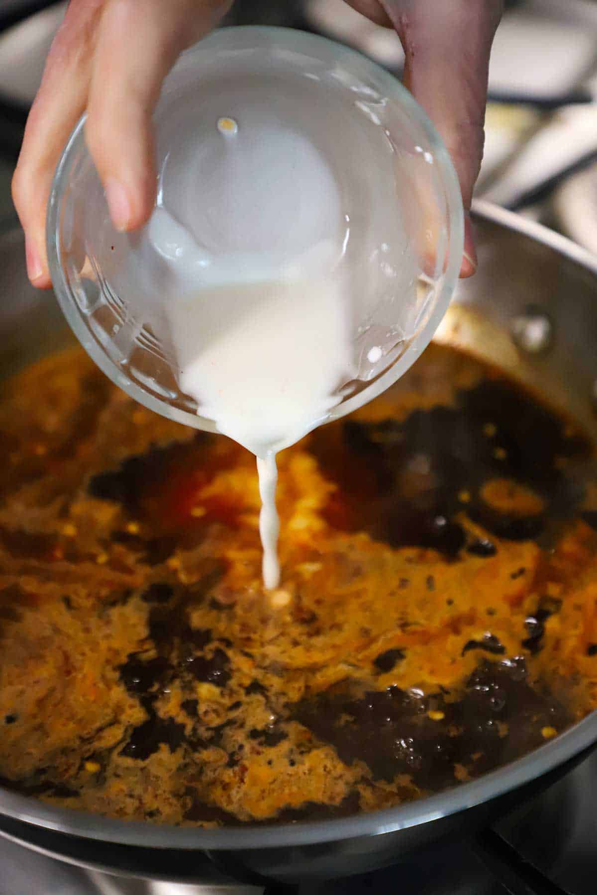 A person pouring a cornstarch slurry from a small glass bowl into a stainless silver skillet filled with a simmering Asian sauce.