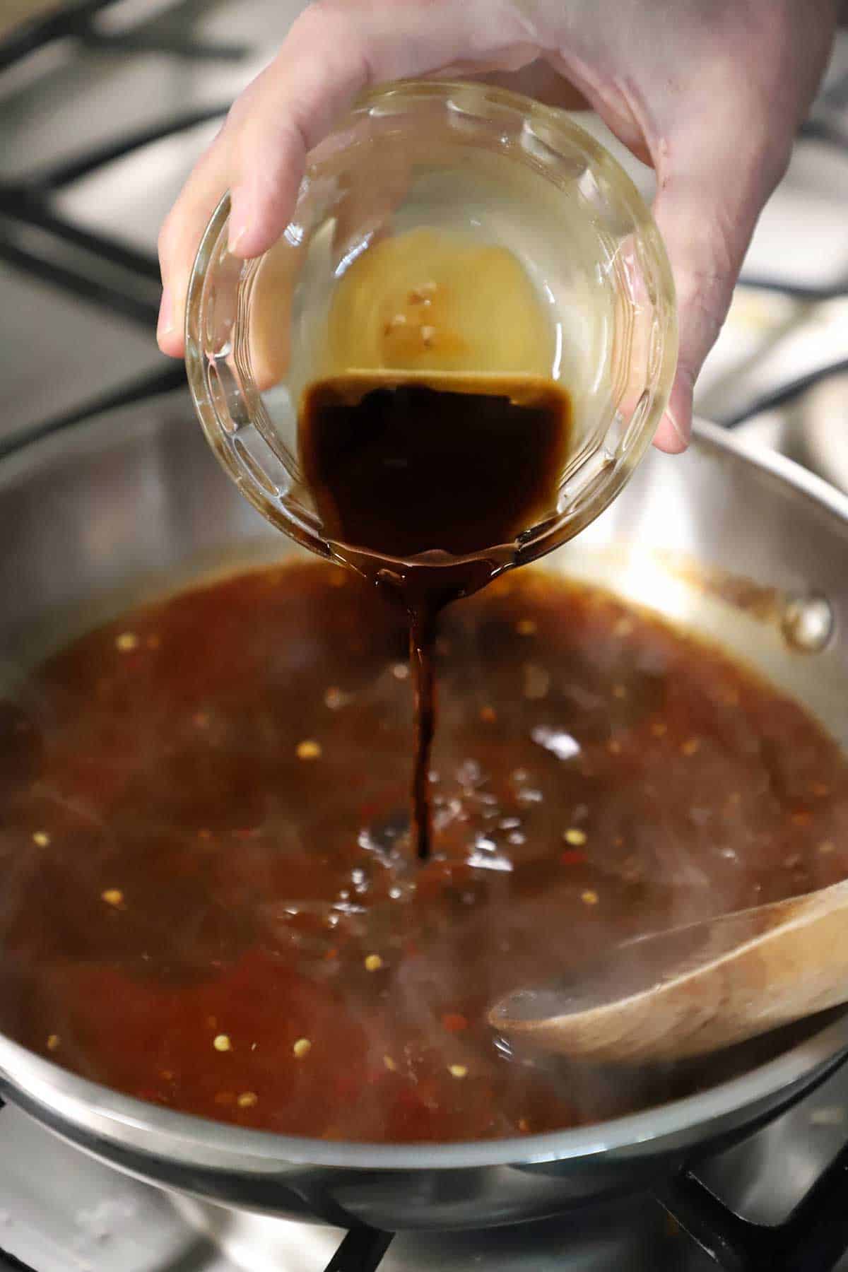 A person pouring soy sauce into a small skillet filled with a simmering Asian garlic chili sauce on a gas stove.