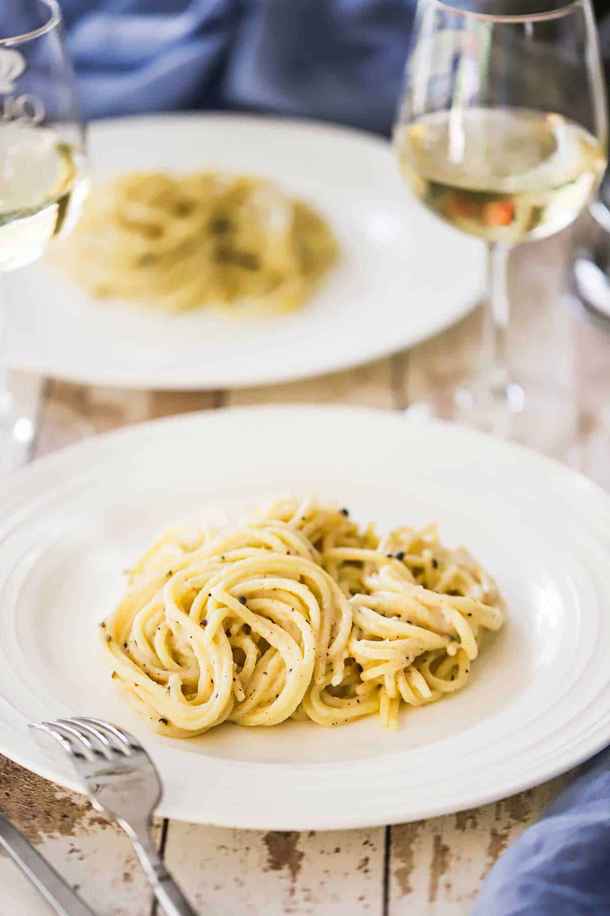 A white dinner plate filled with a serving of tonnarelli cacio e pepe with a glass of white wine nearby. 