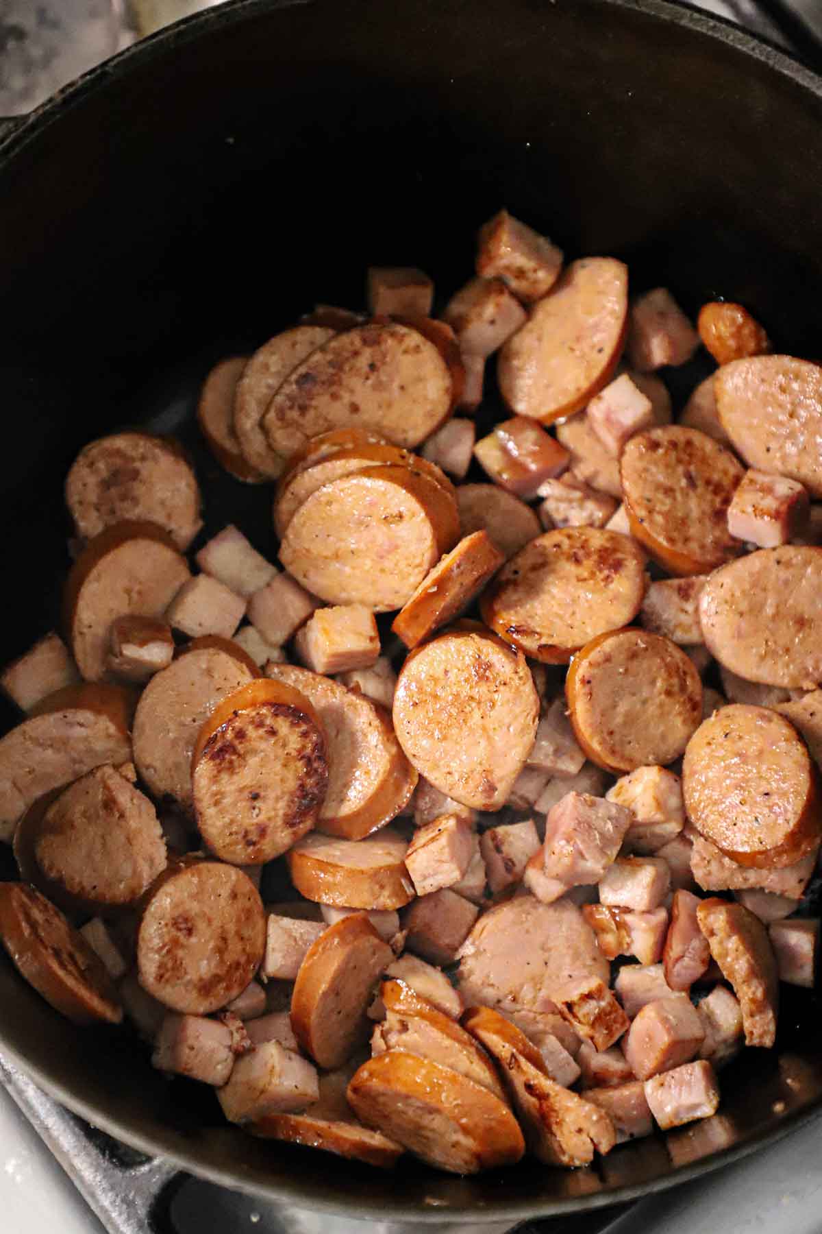 An overhead view of sliced andouille sausage and cubed smoked ham that has been browned in a large black pot.