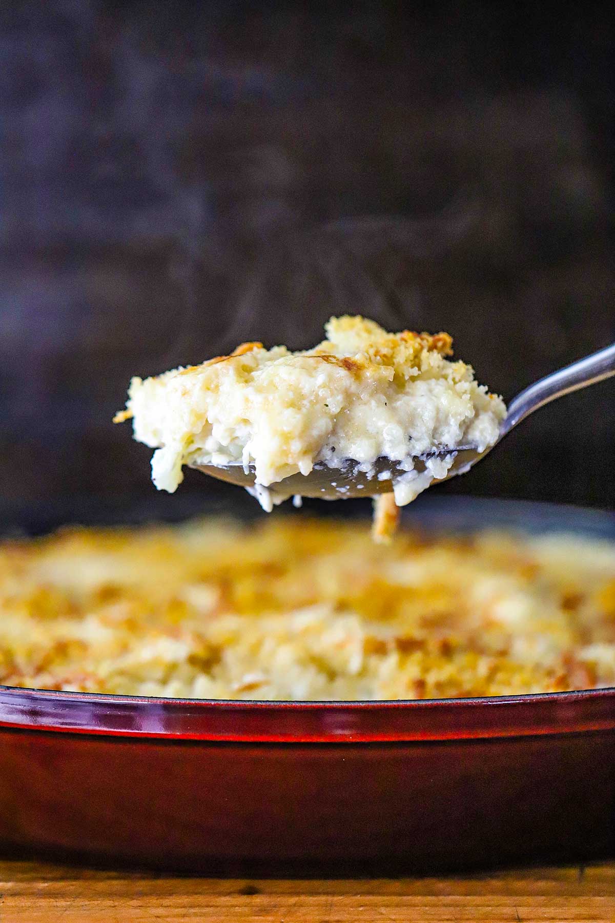A serving spoon being raised from a baking dish holding a steaming serving of creamy cauliflower gratin. 