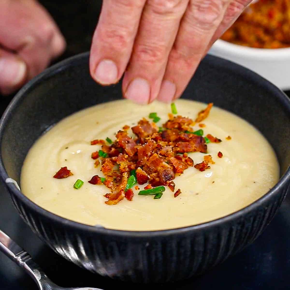 A person sprinkling crumbled cooked bacon onto a bowl filled with creamy Irish potato soup. 