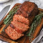 A glazed ham loaf sitting on a small cutting board and has a few slices cut sitting next to sprigs of thyme and rosemary.