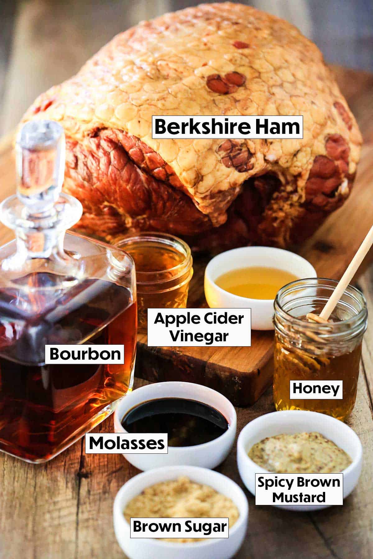 An arrangement of ingredients for honey bourbon glazed ham including a smoked ham, a sniffer bottle of bourbon, vinegar, honey, mustard, and molasses. 