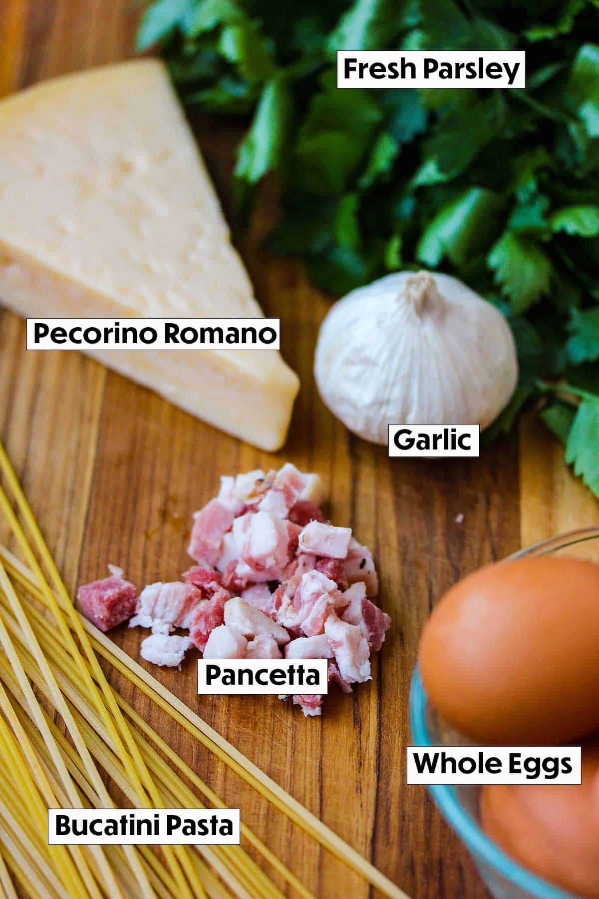 An arrangement of ingredients for pasta carbonara including pecornio romono cheese, parsley, pancetta, pasta, and whole eggs. 