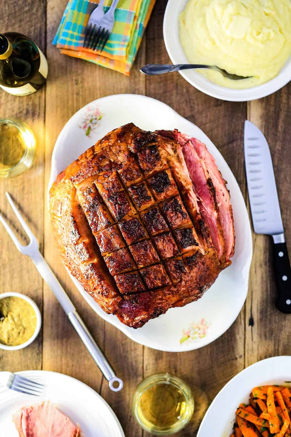 A large Berkshire ham roast that has been scored on top resting on a white oval platter and surrounded by various side dishes. 
