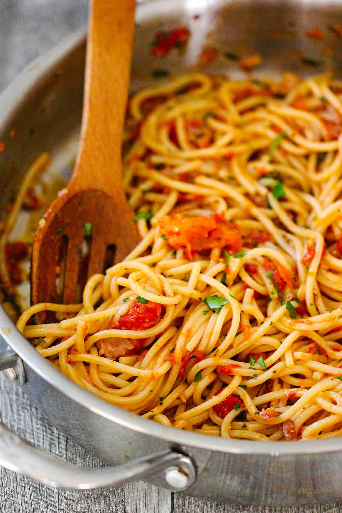 A large stainless skillet filled with pasta pomodor with a wooden slotted spoon stuck in the side. 