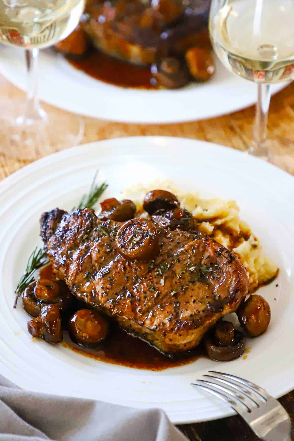 A serving of pork chop agrodolce with mushrooms on a white dinner plate with a glass of white wine next to it. 