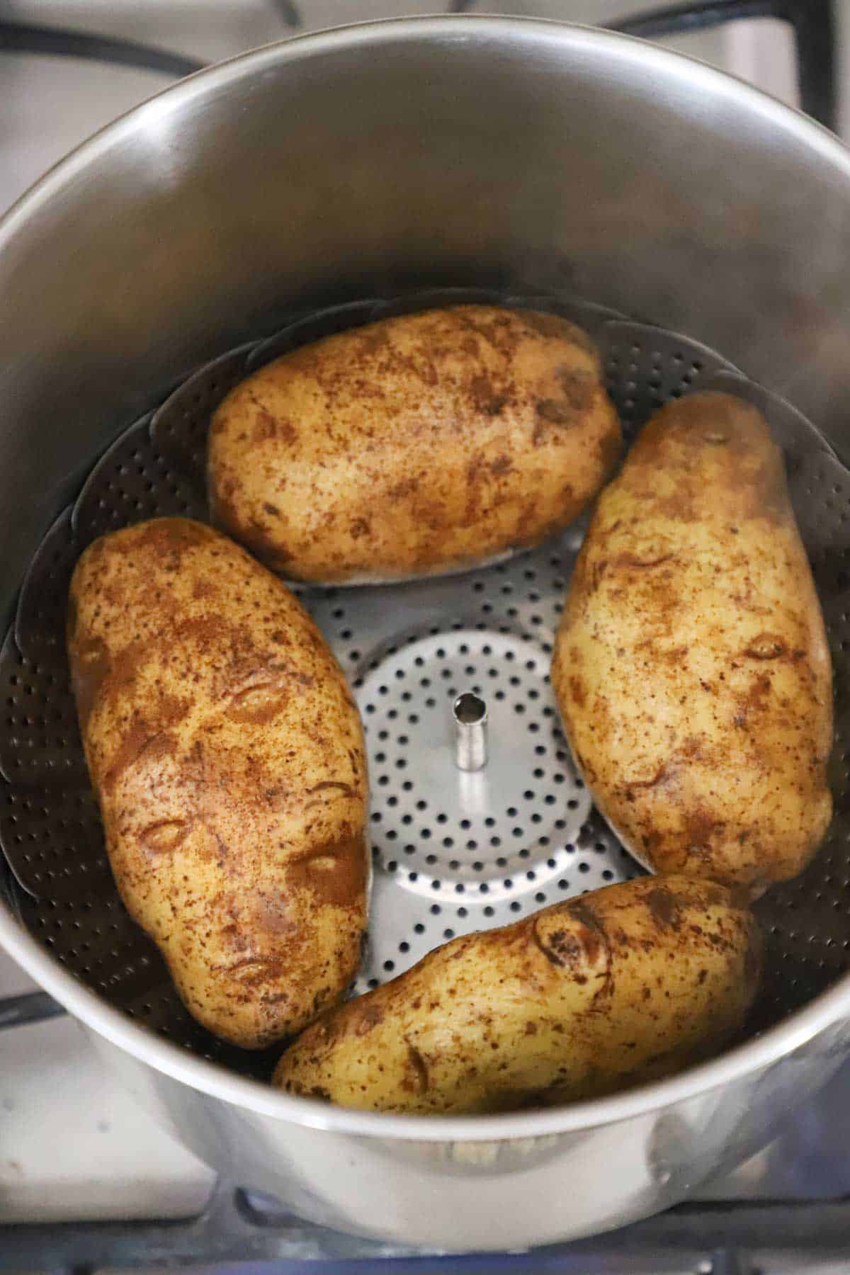 An overhead view of four large Russet potatoes resting inside a stock pot that is lined with a steamer basket in the bottom underneath the potatoes.