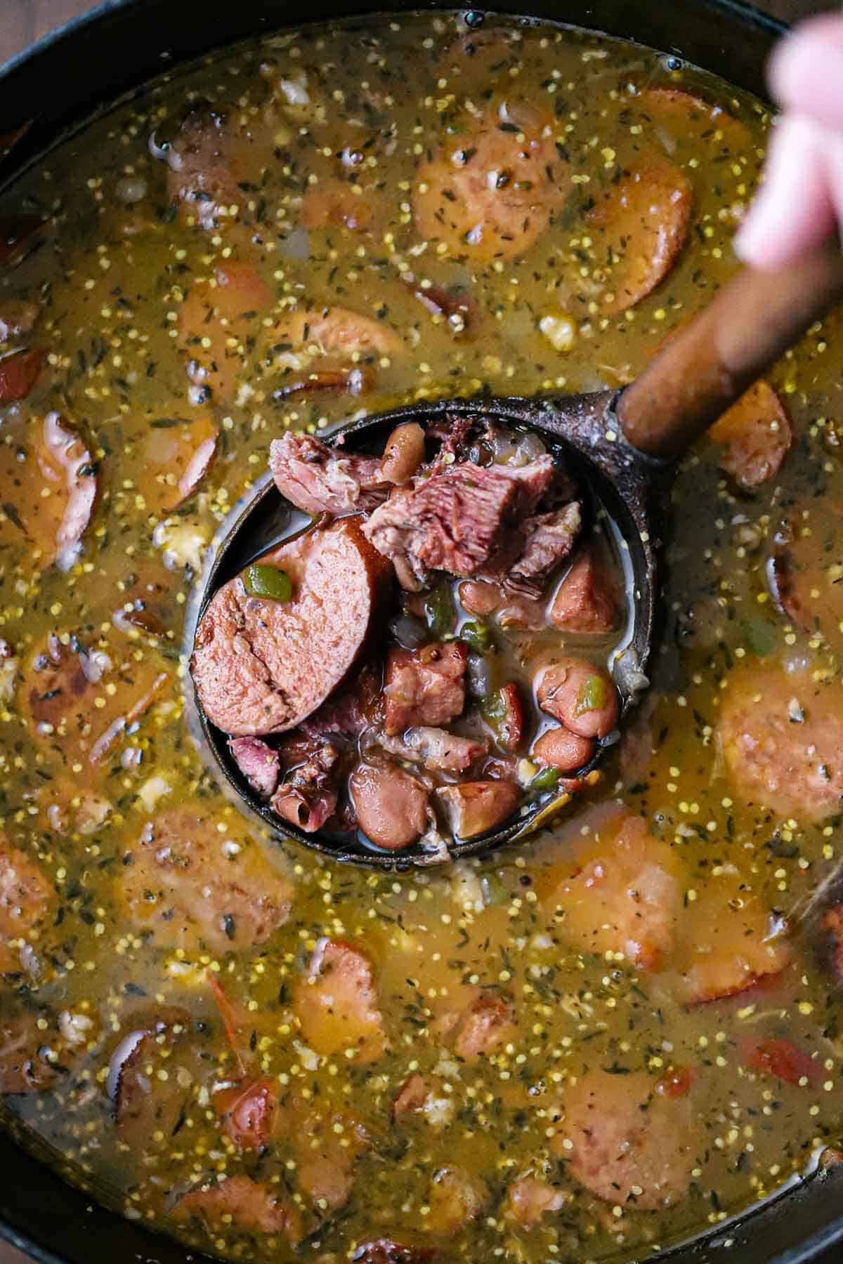 An overhead view of a person lifting a large wooden ladle out of a pot filled with a thick broth, course-ground mustard, red kidney beans, and smoked sausage and ham.