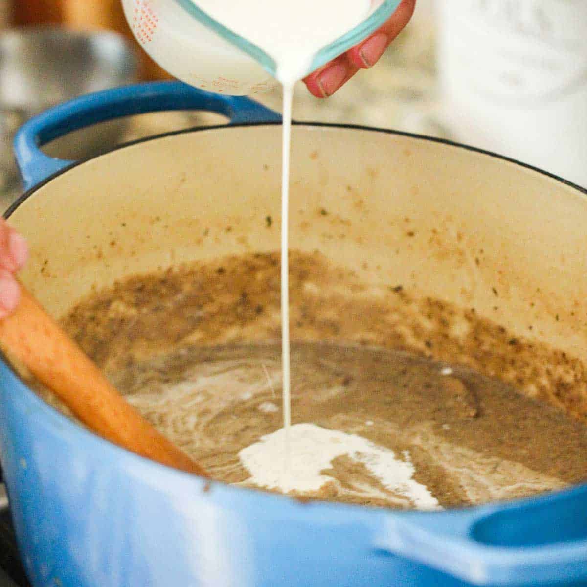 A person pouring cream into an oval Dutch oven that has simmering mushroom soup in it. 