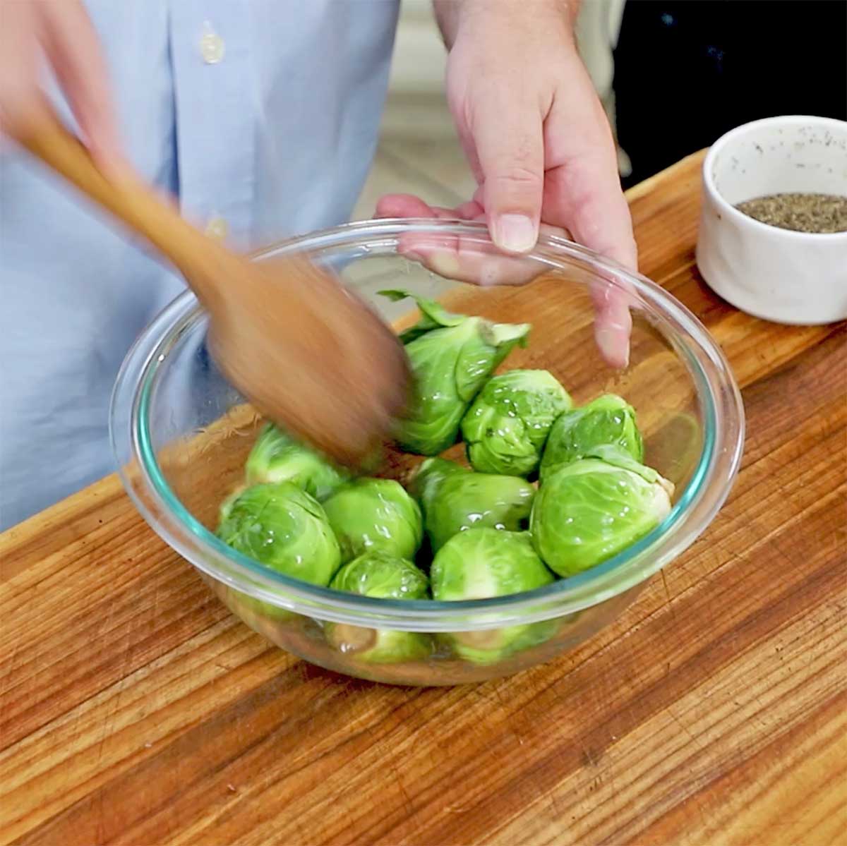 A person using a wooden spoon to toss whole Brussels sprouts with olive oil in a glass bow. 