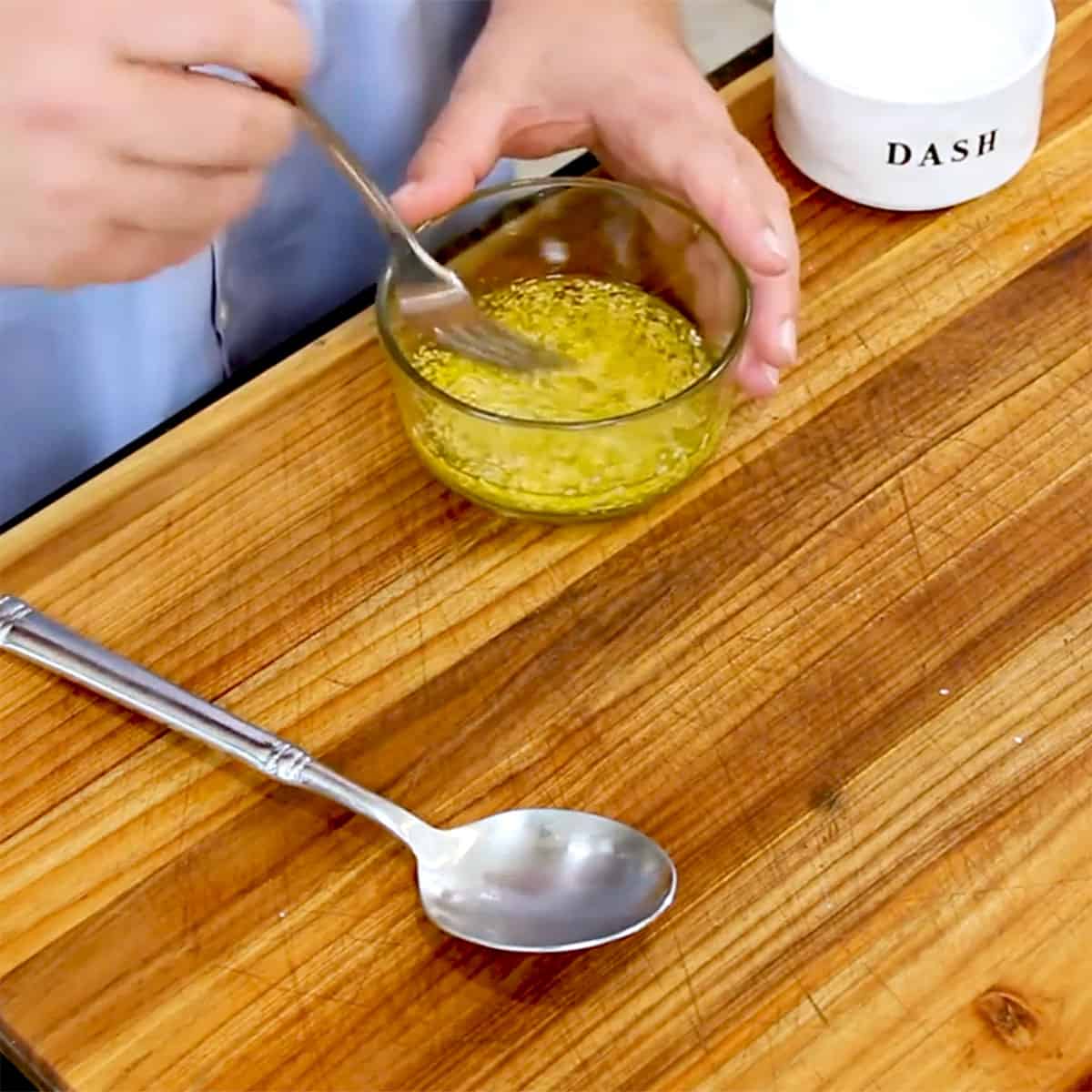 A person using a fork to stir a lemon sesame seed vinaigrette in a small glass bowl. 