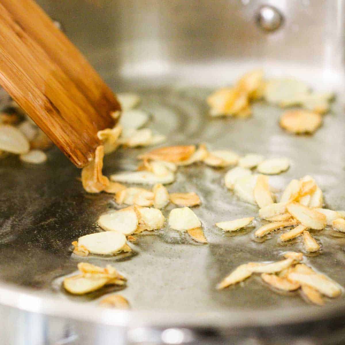 Slices of garlic being sautéed in oil in a large stainless steel skillet with a wooden spatula in the skillet. 