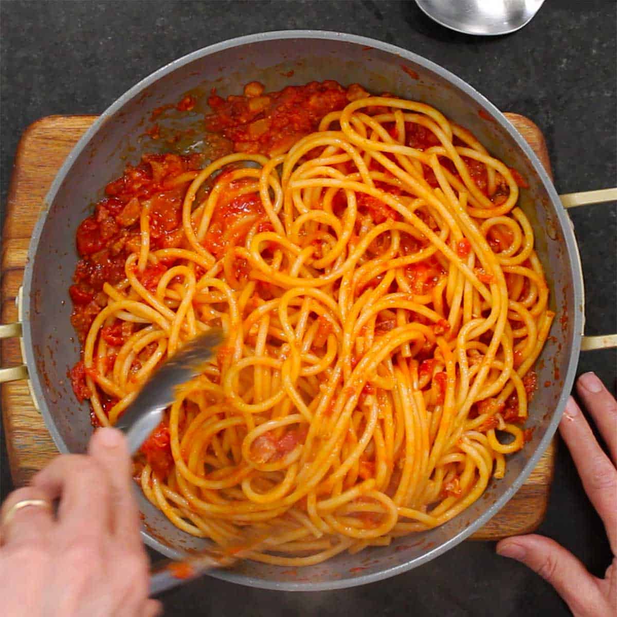 A person using a pair of metal tongs to toss cooked pasta in an Amatriciana sauce. 