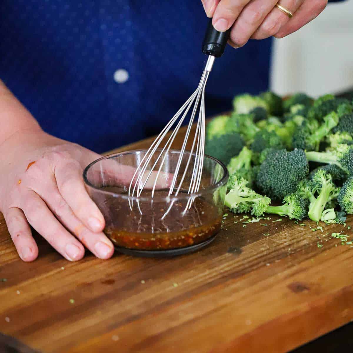 A person using a whisk to mix chili sauce, soy sauce, and lemon juice in a small bowl sitting next to a pile of broccoli florets. 