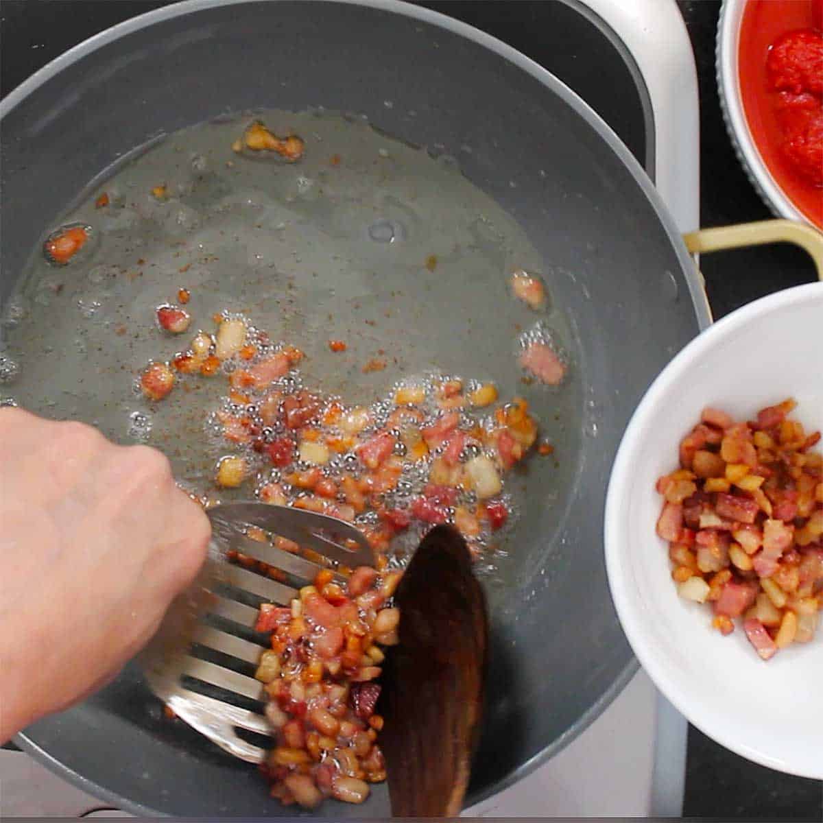 A person using a metal spatula to remove cripsy guanciale from a hot skillet. 