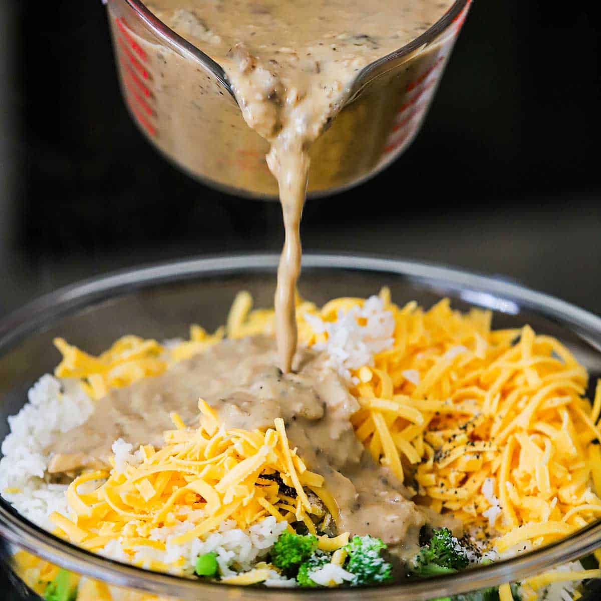A person pouring homemade cream of mushroom soup into a bowl filled with shredded cheddar, broccoli florets, and steamed white rice. 