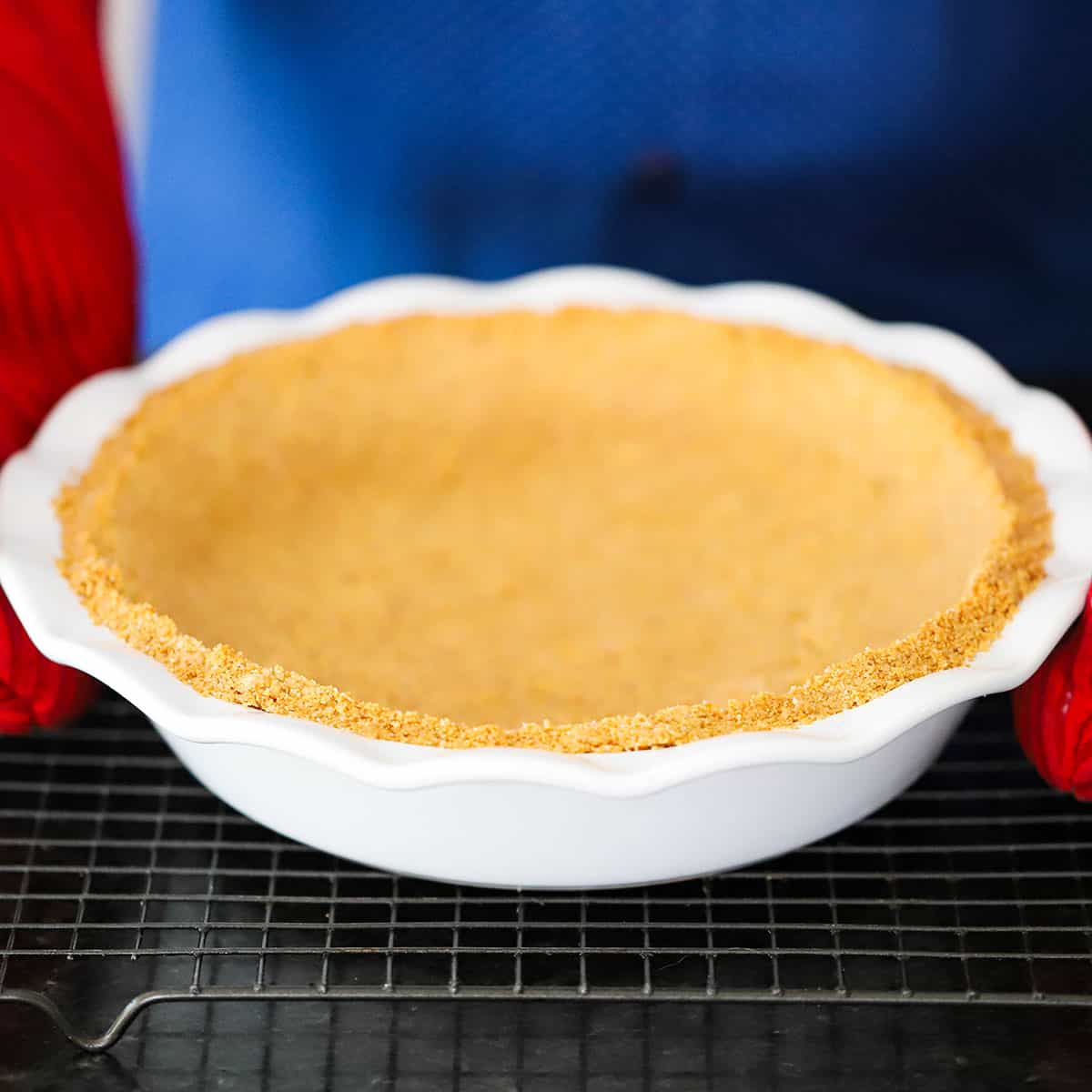 A person holding a white pie dish that has a graham cracker crust that has just come out of the oven. 