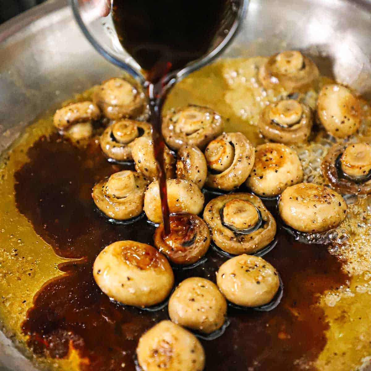 Balsamic vinegar being poured from a glass cup into a skillet filled with honey and sautéed whole mushrooms. 