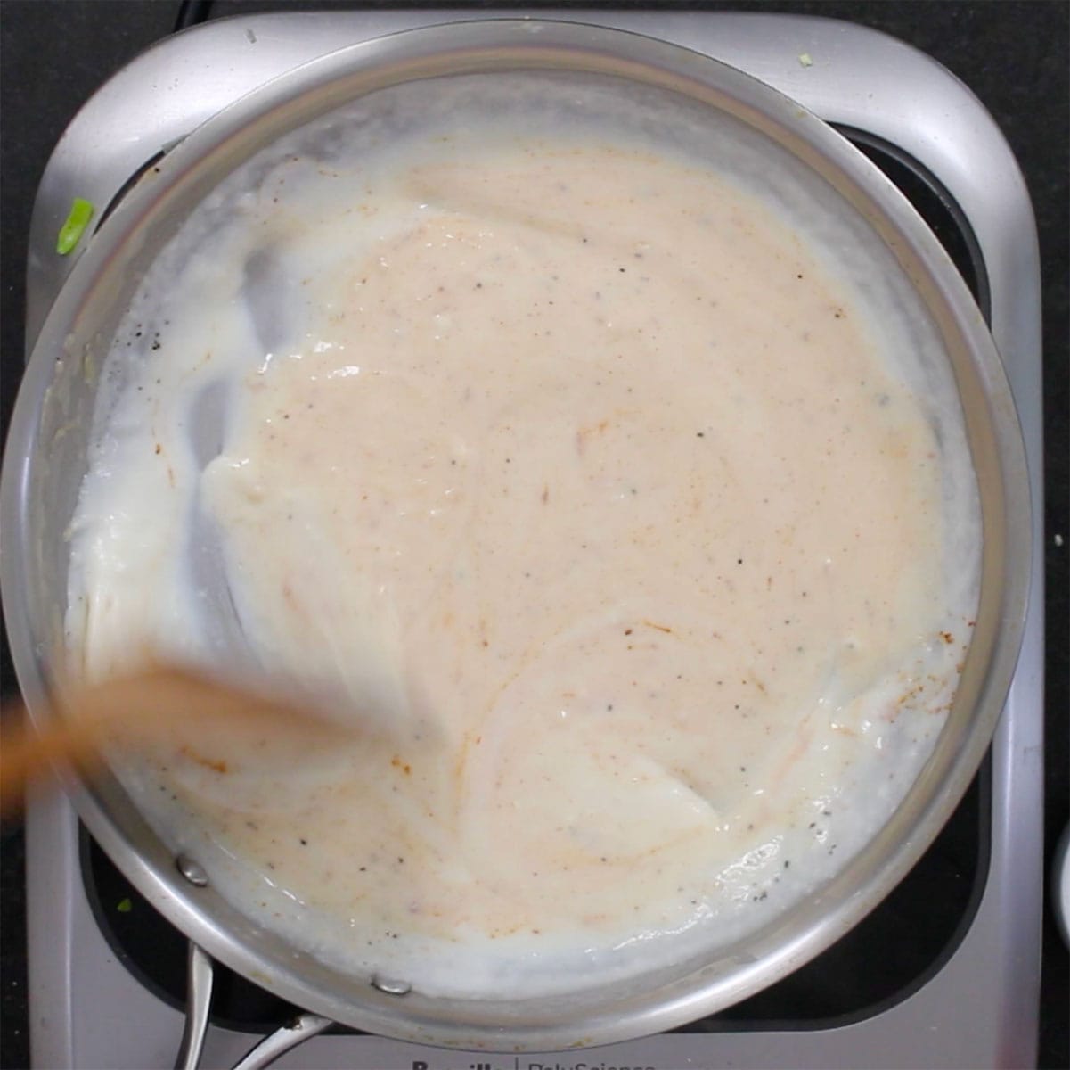 A wooden spatula being used to stir cream that has been incorporated into a white roux in a skillet. 