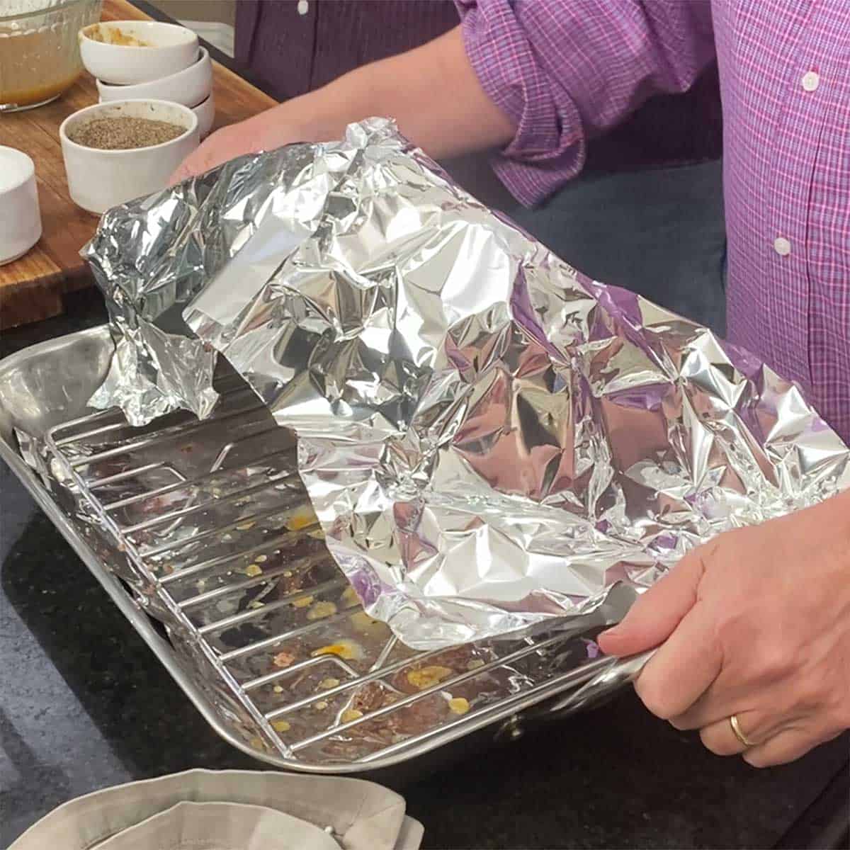 A person lifting a roasting pan that has a foil-covered ham in it. 