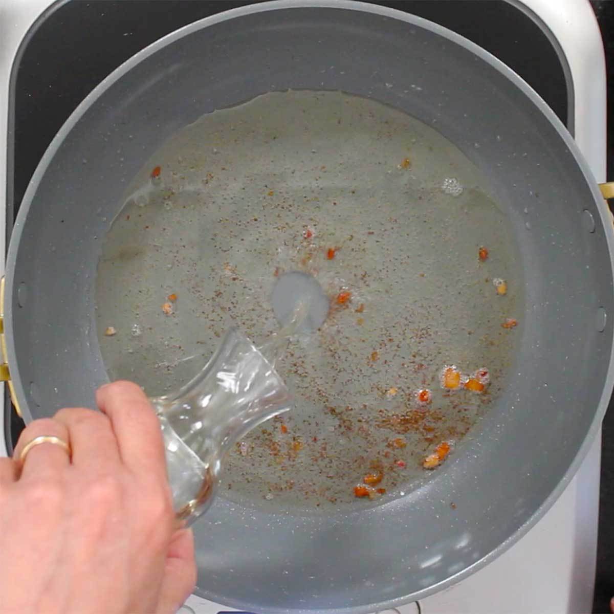 A person pouring white wine from a small carafe into a skillet with simmer pork grease. 
