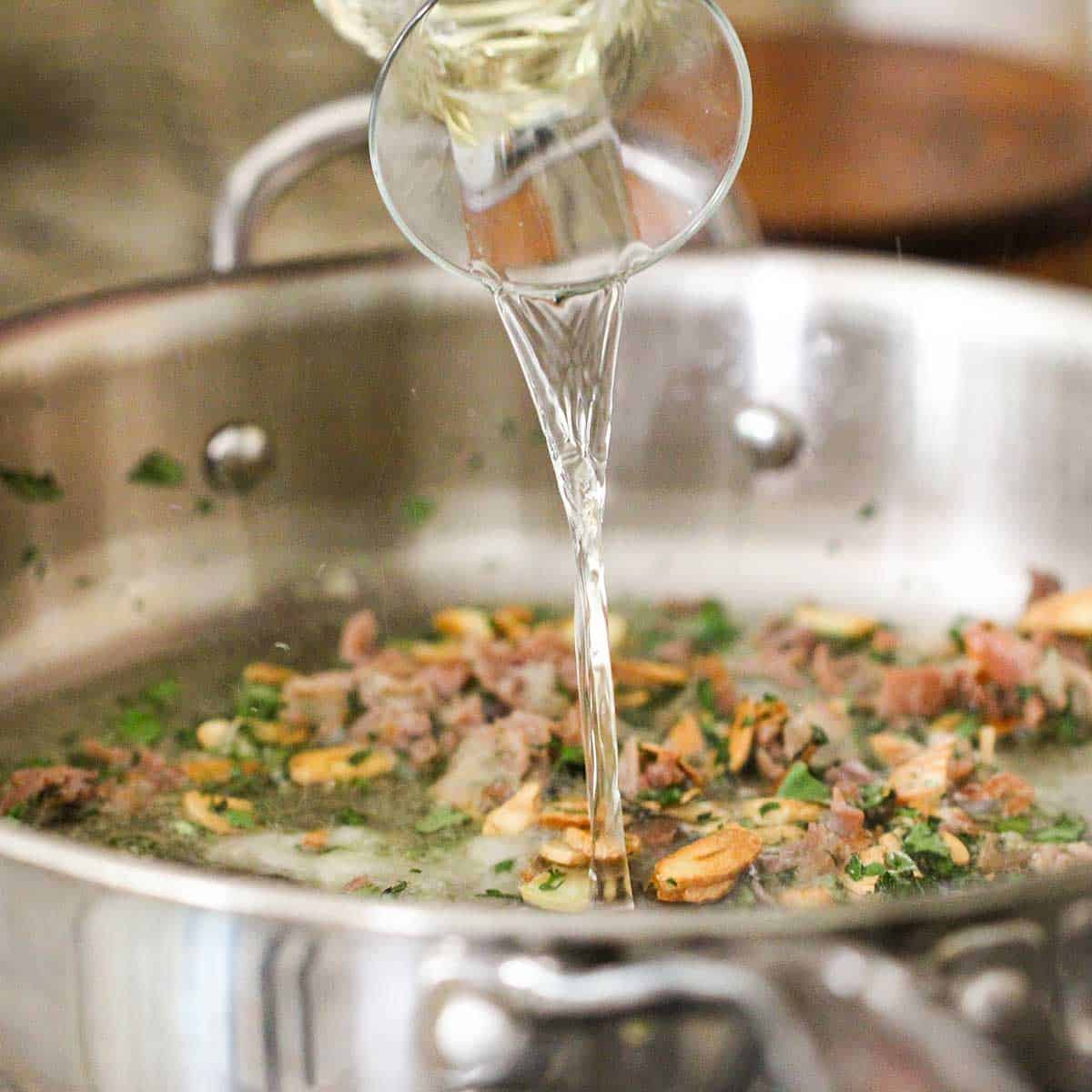 White wine being poured from a small carafe into a skillet filled with sautéed garlic, prosciutto, and herbs. 