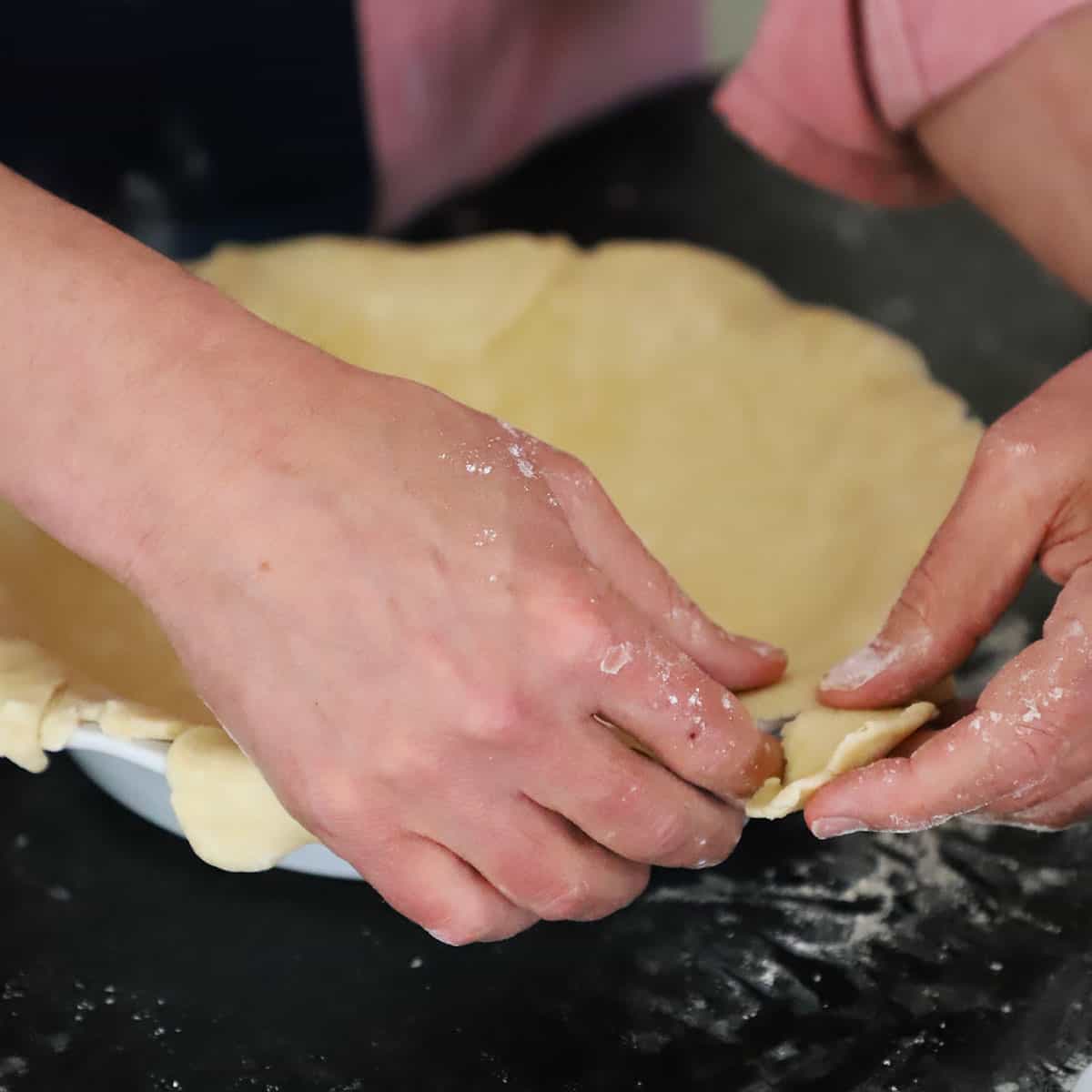 A person trimming away excess pie dough from dough that has been fitted into a pie dish. 