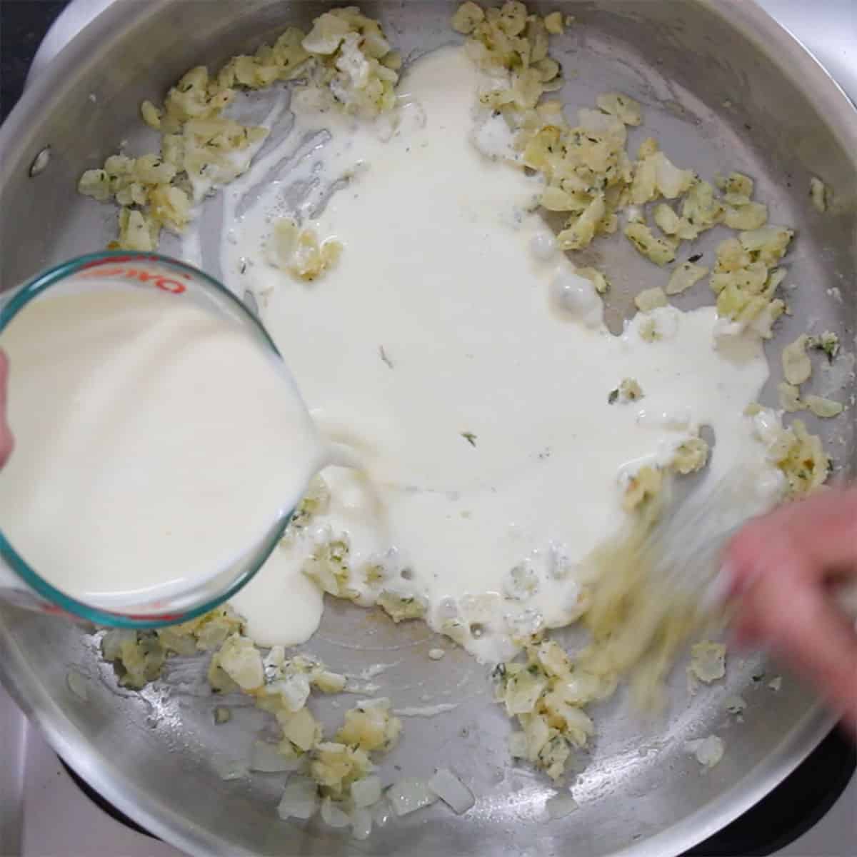 A person pour cream into a skillet that has sautéed onions and flour in it. 