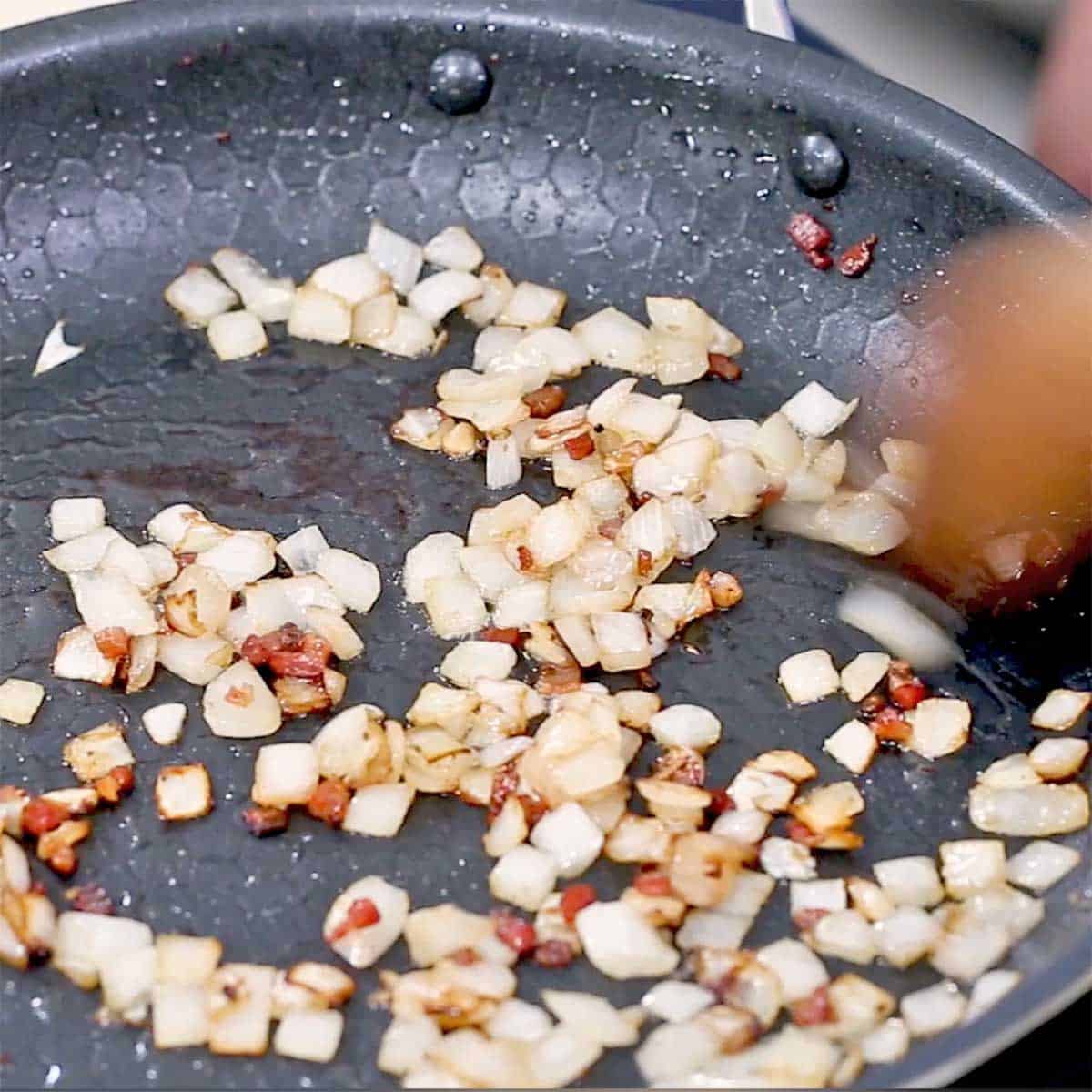 A person using a wooden spatula to sauté chopped onions in a skillet with cooked pancetta. 