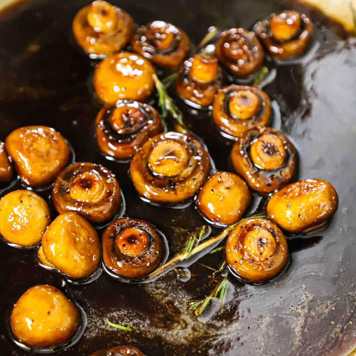 Whole sautéed mushrooms simmering in an agrodolce sauce with a sprig of rosemary also in the sauce. 