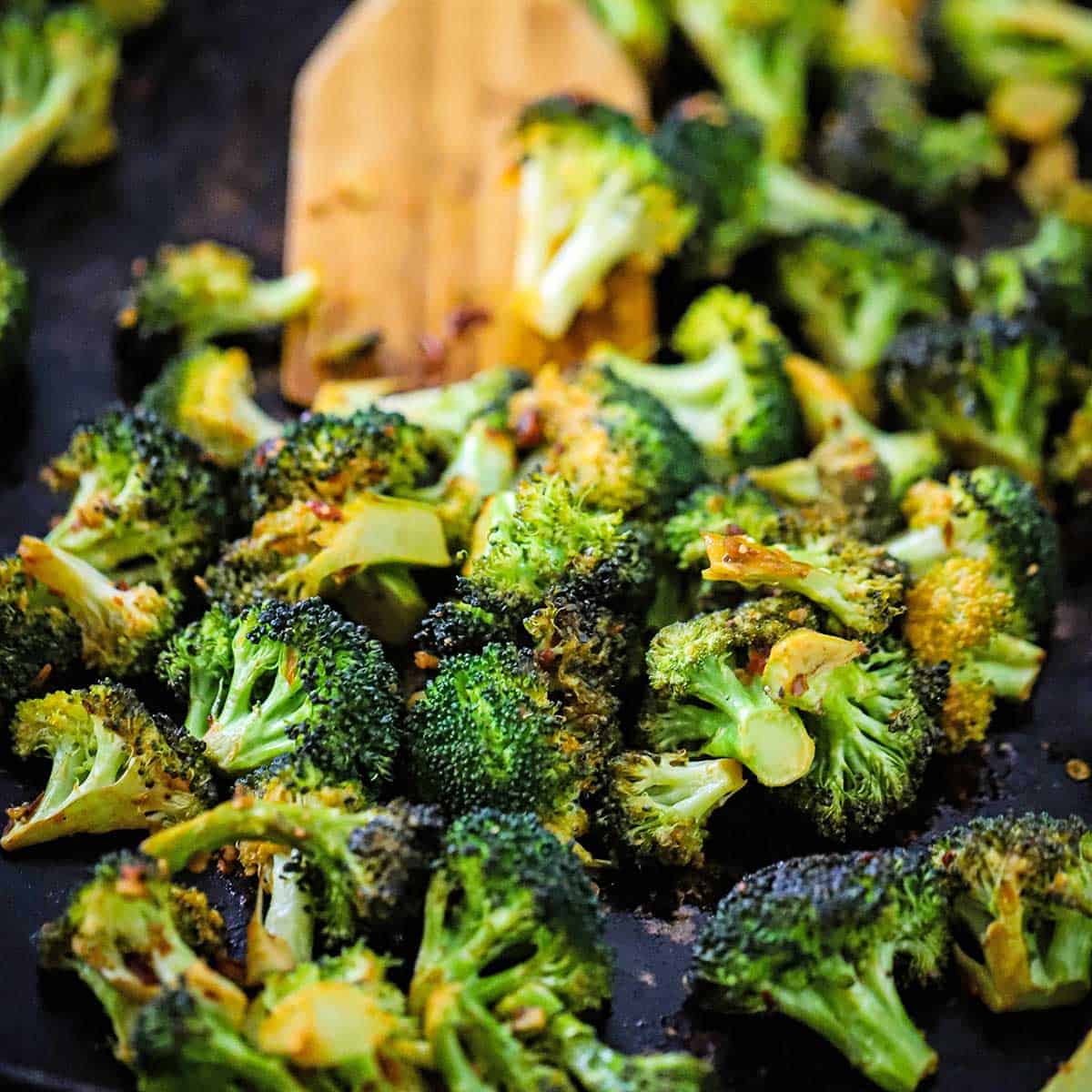 Roasted broccoli with a kick on a baking sheet with a wooden spatula inserted in the middle of the veggies. 