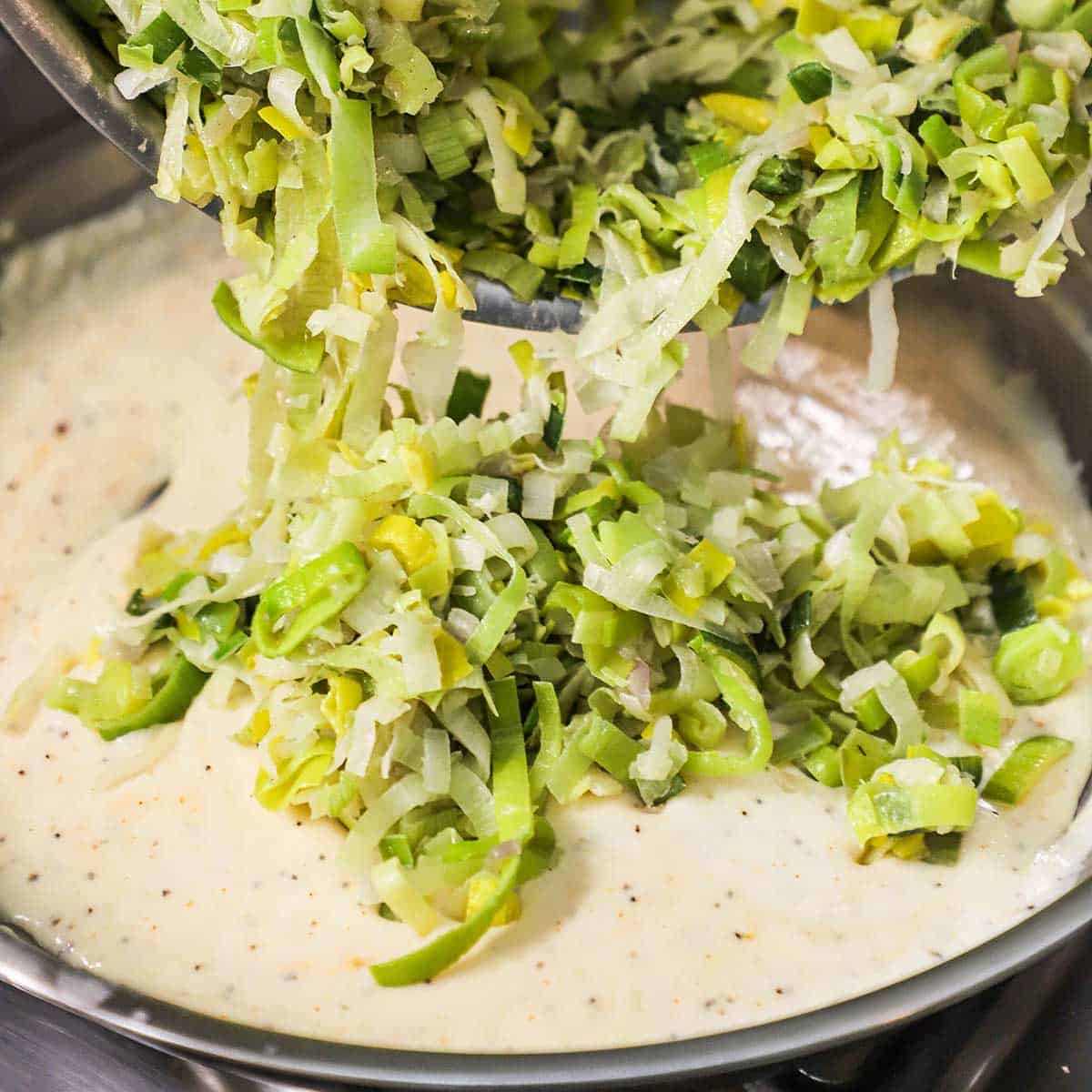 Sautéed leeks and onions being tranfserred into a skillet filled with a creamy sauce. 