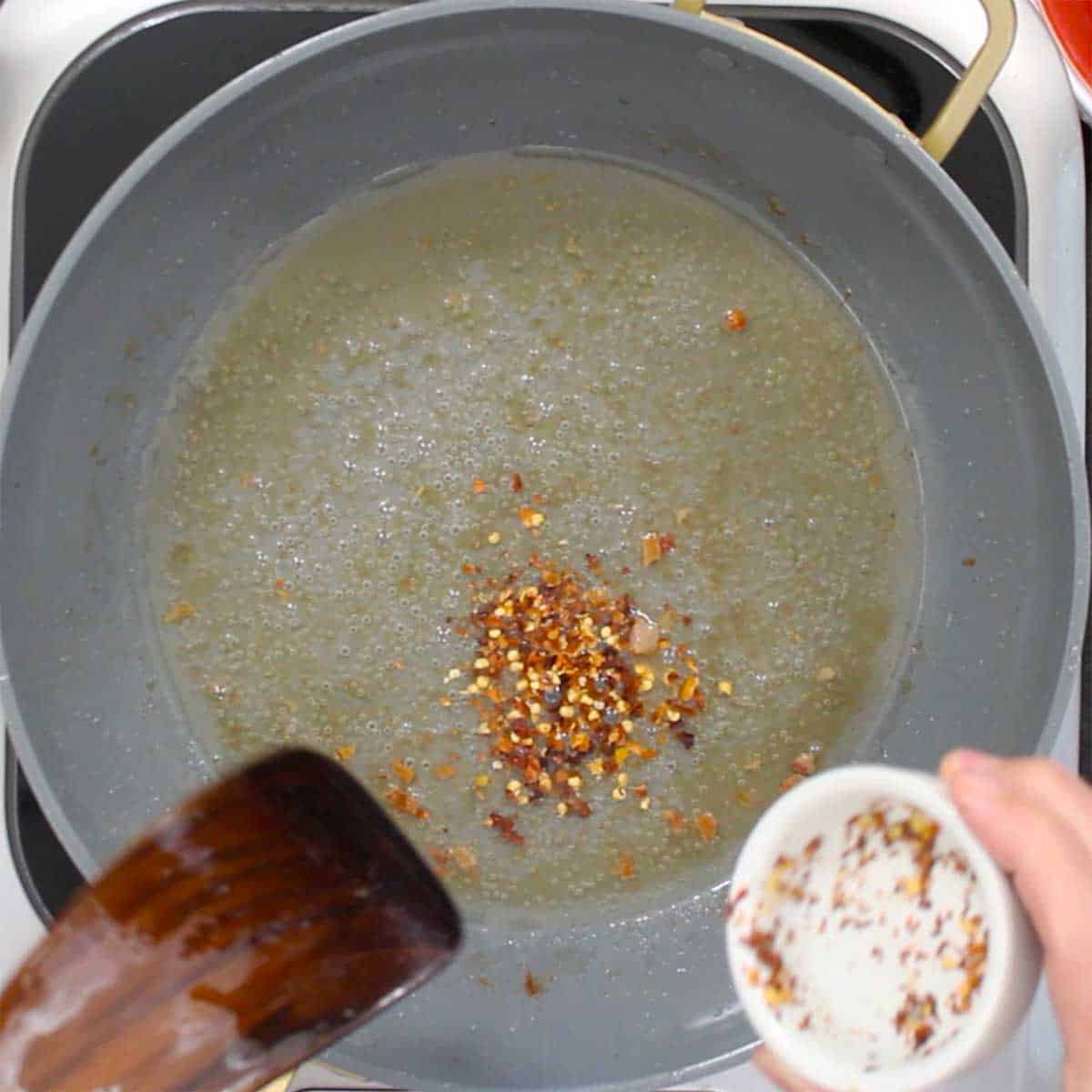 A person dumping red pepper flakes into a skillet filled with simmering white wine and pork grease. 