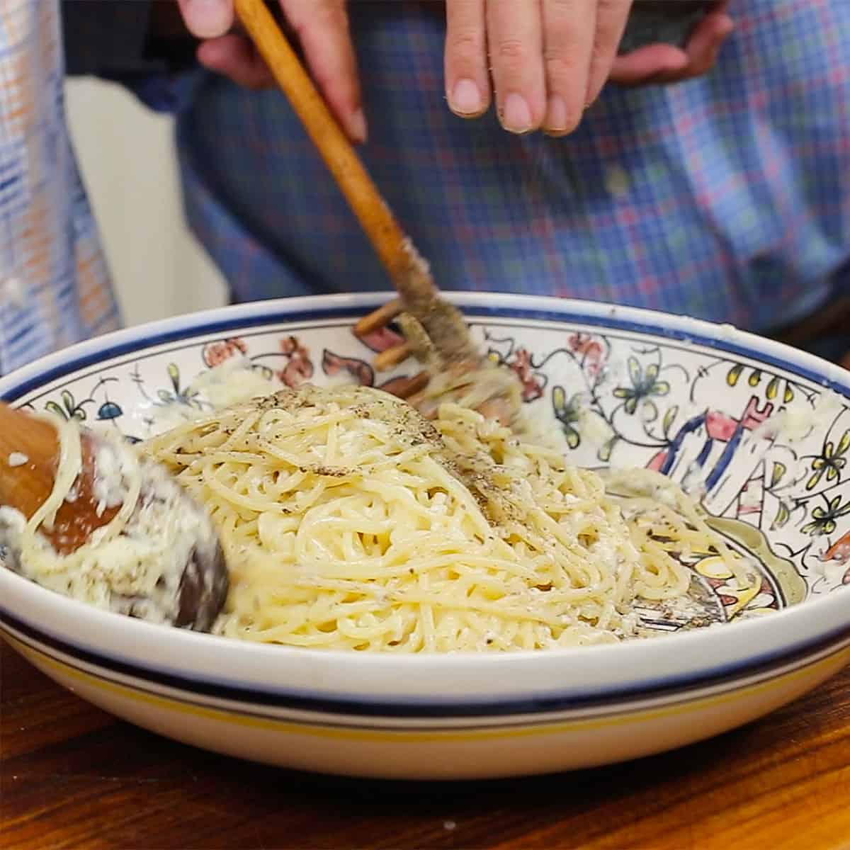 A person sprinkling ground black peppercorns over the top of cooked pasta in a cheese sauce in a large pasta bowl. 