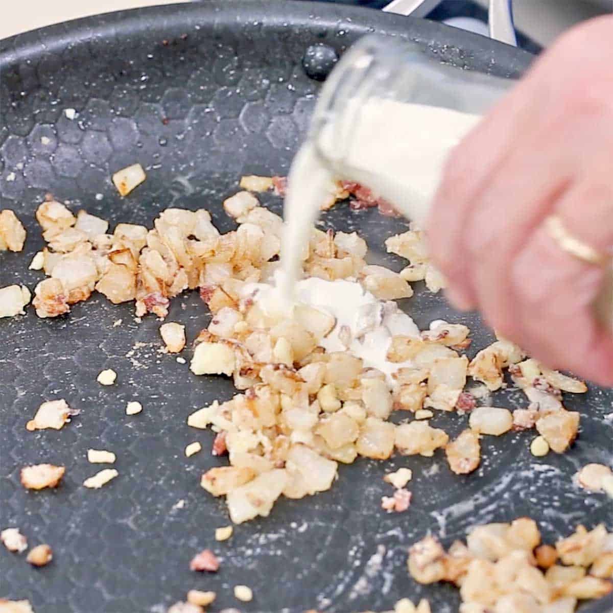 A person pouring cream from a glass jug into a skillet with sautéed onions, pancetta, and cooked flour. 